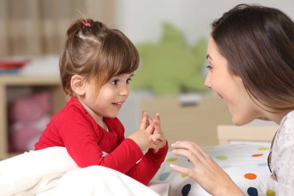 Sprachentwicklung Ein kleines Mädchen in einem roten Oberteil sitzt fröhlich unter einer weißen Decke und unterhält sich lebhaft mit einer lächelnden Frau.
