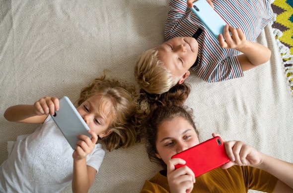 Medienkosum Drei Kinder liegen entspannt auf dem Rücken auf einem Teppich und schauen konzentriert auf ihre Smartphones. Jedes hält ein eigenes Gerät in der Hand, während sie nebeneinander liegen.