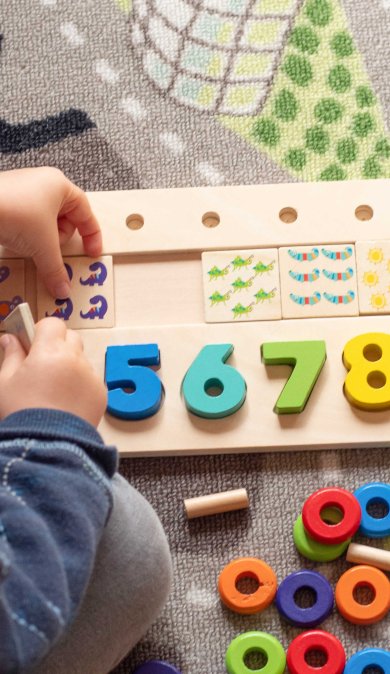 Ein Kind sitzt auf einem bunten Teppich und spielt mit einem Holzpuzzle aus bunten Zahlen und Symbolen.