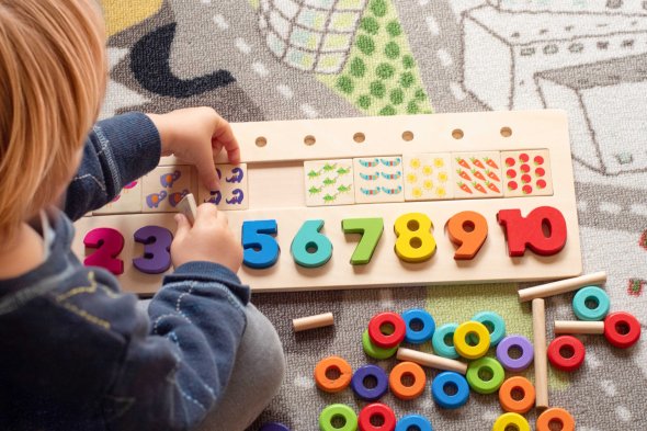 Zahlen und Mengen Ein Kind sitzt auf einem bunten Teppich und spielt mit einem Holzpuzzle aus bunten Zahlen und Symbolen.