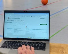 Ein Laptop auf dem Hallenboden einer Sporthalle zeigt die Online-Buchungsplattform des Landkreises Hersfeld-Rotenburg für kreiseigene Sportanlagen. Im Hintergrund liegt ein Basketball.