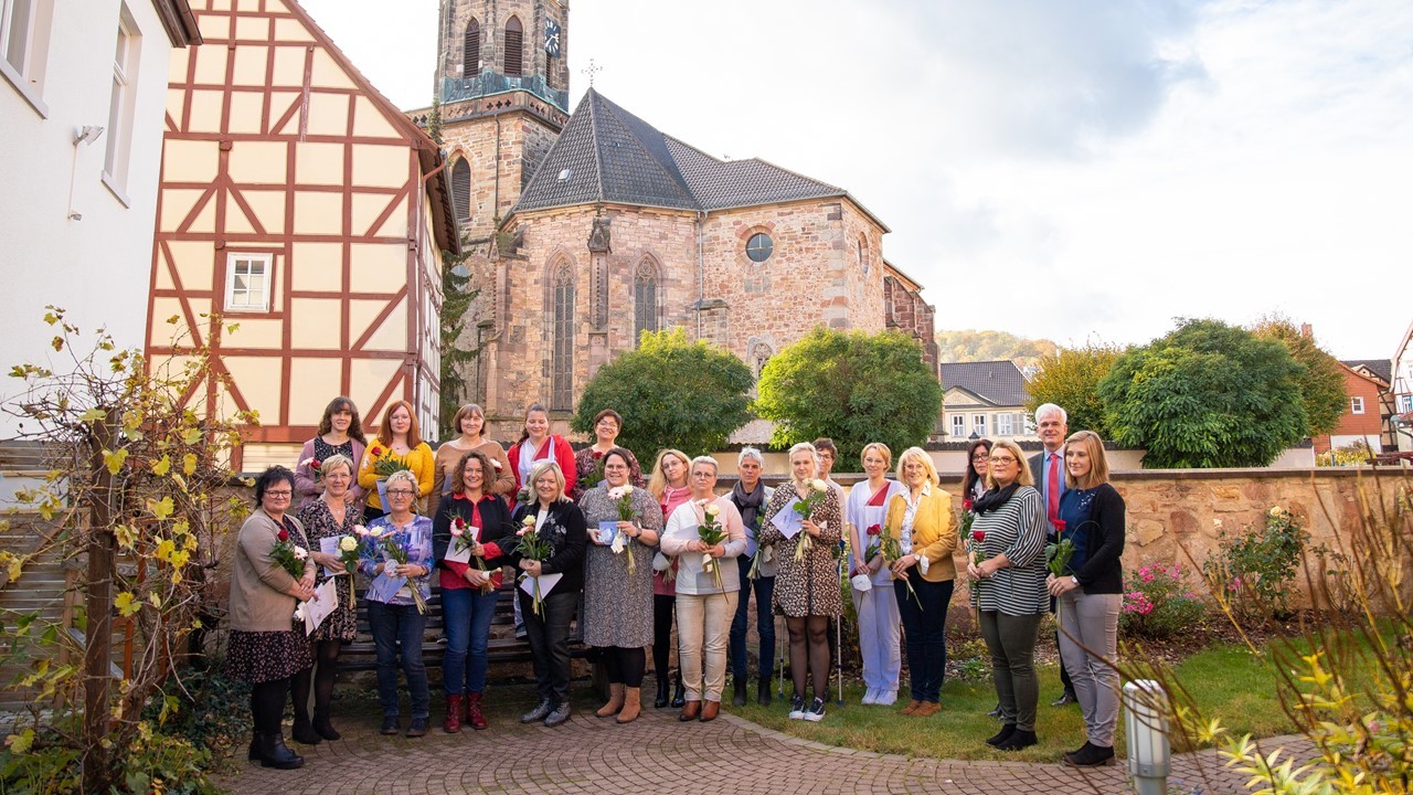 Eine Menschengruppe hauptsächlich aus geehrten Frauen, die jeweils eine Rose und Urkunde in der Hand halten, steht für ein Gruppenfoto vor einer Mauer in einem Garten. Im Hintergrund ist die Kirche von Rotenburg zu erkennen.