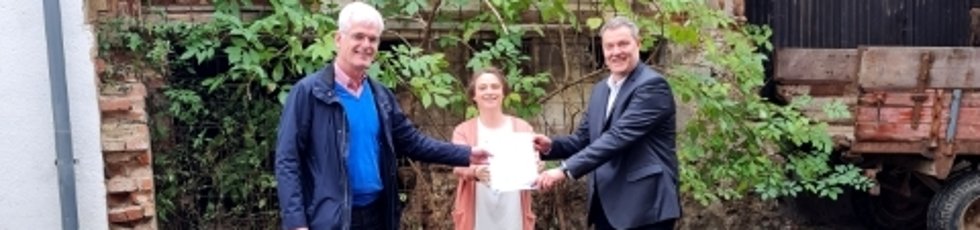 Landrat Torsten Warnecke (links) überreicht den Förderbescheid für den Beratungsvertrag an Alheims Bürgermeister Jochen Schmidt, zwischen den beiden steht SPD-Landtagsabgeordnete Karina Fissmann. Die drei stehen vor einer alten Fachwerkruine. 