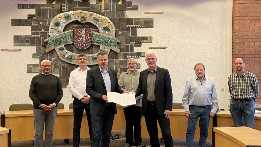 Landrat Torsten Warnecke (vorne rechts) übergibt Bebras Bürgermeister Stefan Knoche einen Förderbescheid. Im Hintergrund zu sehen sind (von links) Stadtrat Morcos Bekheit, Erster Stadtrat Volkmar Hanf (beide Gemeinsam für Bebra) sowie die beiden Stadträte Horst Taube (CDU) und Bernd Schill (SPD) sowie Markus Knobl vom Bau- und Planungsamt der Stadt Bebra. Das Foto ist im Sitzungssaal der Stadt Bebra aufgenommen, im Hintergrund an der Wappen ist das Wappen der Stadt Bebra zu erkenn.