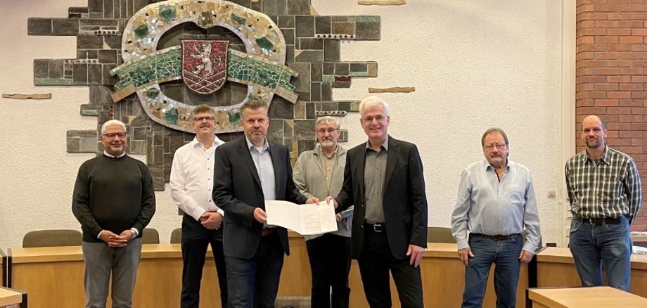 Landrat Torsten Warnecke (vorne rechts) übergibt Bebras Bürgermeister Stefan Knoche einen Förderbescheid. Im Hintergrund zu sehen sind (von links) Stadtrat Morcos Bekheit, Erster Stadtrat Volkmar Hanf (beide Gemeinsam für Bebra) sowie die beiden Stadträte Horst Taube (CDU) und Bernd Schill (SPD) sowie Markus Knobl vom Bau- und Planungsamt der Stadt Bebra. Das Foto ist im Sitzungssaal der Stadt Bebra aufgenommen, im Hintergrund an der Wappen ist das Wappen der Stadt Bebra zu erkenn.