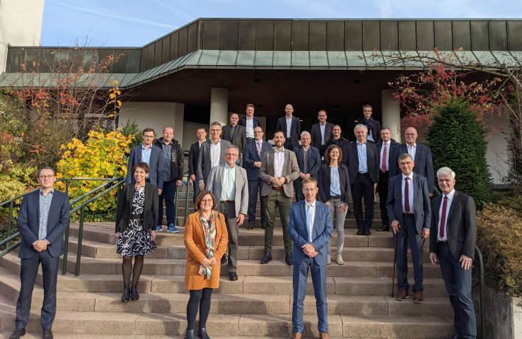 Eine Menschengruppe steht mit Abstand zueinander auf einer Treppe für ein Gruppenbild. Rechts und links sind Sträucher mit bunt gefärbten Blättern zu sehen, im Hintergrund der Eingang einer Veranstaltungshalle.