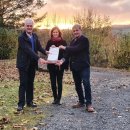 Landrat Torsten Warnecke (links) übergibt Katrin Anders, der Geschäftsführerin des Zweckverbands Knüll, und Walter Glänzer, Neuensteins Bürgermeister und Vorsitzender des Zweckverbands Knüll, einen Förderbescheid. Die drei stehen auf einem Schotterweg, der mit Laub bedeckt ist. Im Hintergrund sind bunte Herbststräucher sowie dahinter Felder zu erkennen. Der Himmel leuchtet gelb-orange im Sonnenuntergang.