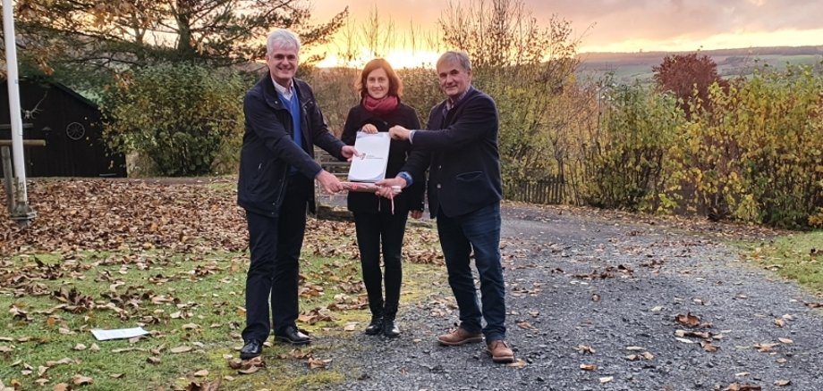 Landrat Torsten Warnecke (links) übergibt Katrin Anders, der Geschäftsführerin des Zweckverbands Knüll, und Walter Glänzer, Neuensteins Bürgermeister und Vorsitzender des Zweckverbands Knüll, einen Förderbescheid. Die drei stehen auf einem Schotterweg, der mit Laub bedeckt ist. Im Hintergrund sind bunte Herbststräucher sowie dahinter Felder zu erkennen. Der Himmel leuchtet gelb-orange im Sonnenuntergang.