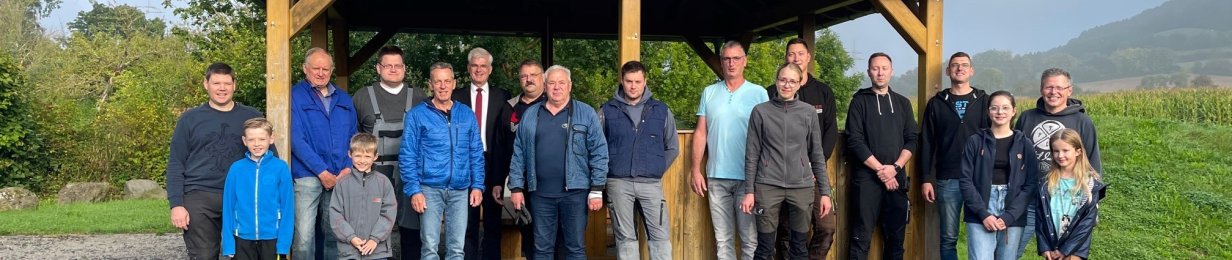 Eine Menschengruppe hat sich für ein Gruppenbild vor einem Holzpavillon mit Blechdach für ein Foto aufgestellt. Zum Pavillon führt ein Schotterweg. Rechts und links ist Rasen, im Hintergrund sind links Bäume und rechts Felder sowie Berge zu erkennen. Vor der Gruppe liegt ein weiß-braun gefleckter Hund.