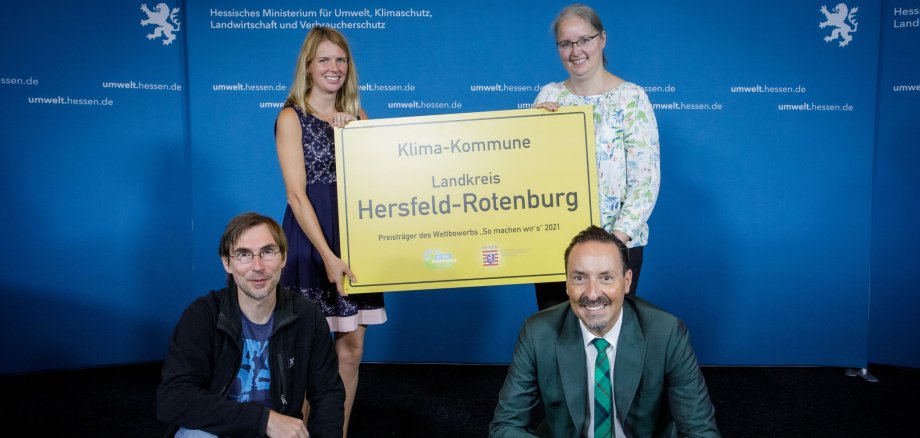 Zwei Frauen stehen vor einer blauen Wand mit dem Löwen-Symbol des Landes Hessen und der weißen Aufschrift Hessisches Ministerium für Umwelt, Klimaschutz, Landwirtschaft und Verbraucherschutz. Die beiden Frauen halten ein gelbes Schild mit der Aufschrift Klima-Kommune Landkries Hersfeld-Rotenburg - Preisträger des Wettbewerbs "So machen wir's" 2021" hoch. Vor den beiden Frauen hocken zwei Männer, einer hat eine Urkunde vor sich aufgestellt.