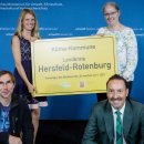 Zwei Frauen stehen vor einer blauen Wand mit dem Löwen-Symbol des Landes Hessen und der weißen Aufschrift Hessisches Ministerium für Umwelt, Klimaschutz, Landwirtschaft und Verbraucherschutz. Die beiden Frauen halten ein gelbes Schild mit der Aufschrift Klima-Kommune Landkries Hersfeld-Rotenburg - Preisträger des Wettbewerbs "So machen wir's" 2021" hoch. Vor den beiden Frauen hocken zwei Männer, einer hat eine Urkunde vor sich aufgestellt.