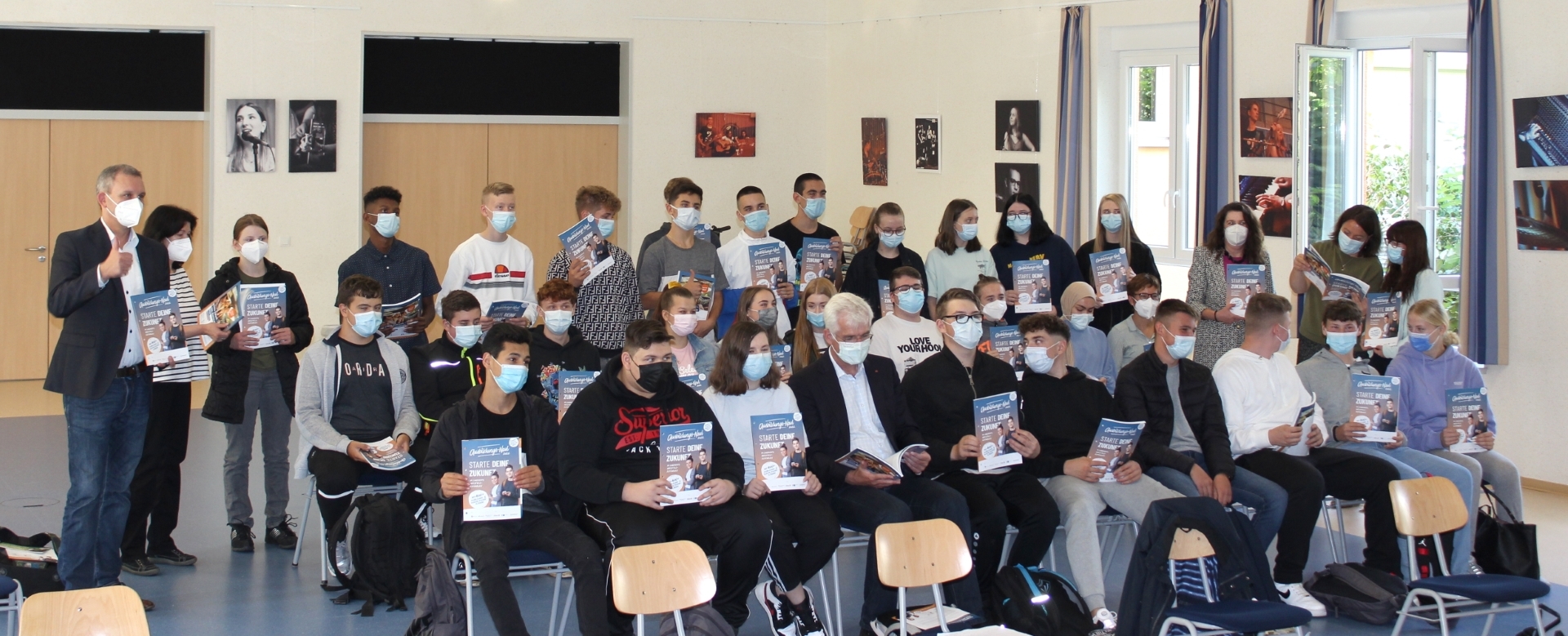 Eine Schulklasse sitzt mit Mundschutzmasken auf auf Stühlen bzw. die letzte Reihe steht und hält eine blaue Broschüre mit der Aufschrift "Ausbildungs-Navi - starte deine Zukunft" hoch.