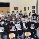 Eine Schulklasse sitzt mit Mundschutzmasken auf auf Stühlen bzw. die letzte Reihe steht und hält eine blaue Broschüre mit der Aufschrift "Ausbildungs-Navi - starte deine Zukunft" hoch. 
