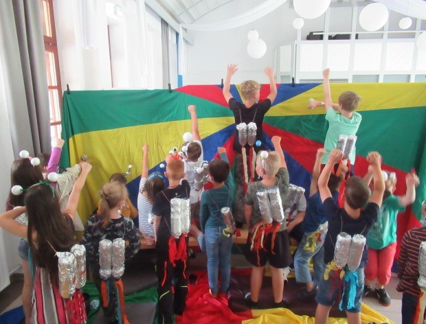 Rückansicht einer Kindergruppe, die alle eine silberne gebastelte Rakete auf ihren Rücken tragen. Die Kids strecken die Hände, als ob sie auf den Mars reisen. Sie stehen vor einer bunten Wand.