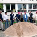 Acht Personen stehen auf einem Schulhof vor einem Berg von Sand. Im Sand liegen Glasfaser-Rohre. Vier Personen tragen einen Helm in der Farbe pink. Drei Personen haben einen Spaten in der Hand. Alle lächeln.