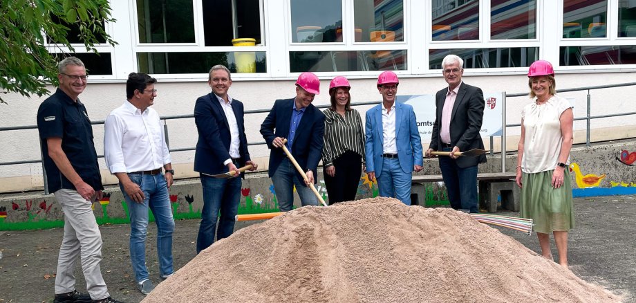 Acht Personen stehen auf einem Schulhof vor einem Berg von Sand. Im Sand liegen Glasfaser-Rohre. Vier Personen tragen einen Helm in der Farbe pink. Drei Personen haben einen Spaten in der Hand. Alle lächeln.