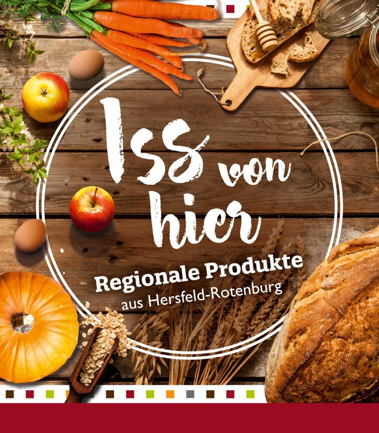 Das Bild zeigt eine Werbegrafik für regionale Produkte aus Hersfeld-Rotenburg. Im Vordergrund sind verschiedene Lebensmittel zu sehen: Möhren, ein Apfel, Eier, ein Stück Brot, ein Glas Honig, ein Kürbis und Haferflocken, die auf einem hölzernen Untergrund arrangiert sind. Über den Lebensmitteln befindet sich der Text "Iss von hier" in großen, weißen, handgeschriebenen Buchstaben, was übersetzt "Essen Sie von hier" bedeutet. Darunter steht "Regionale Produkte aus Hersfeld-Rotenburg" als Hinweis auf die Herkunft der Waren. Das Layout betont die Frische und Natürlichkeit der Produkte und die Unterstützung der lokalen Landwirtschaft. Die Farben und Texturen der Lebensmittel sind reich und ansprechend dargestellt.