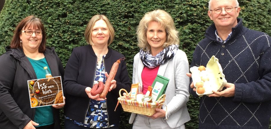 Das Foto zeigt vier Personen, die offenbar eine Kampagne für regionale Produkte unterstützen. Die erste Person links hält ein Schild mit der Aufschrift „Iss von hier“, das wir bereits aus dem vorherigen Bild kennen. Die nächsten zwei Personen halten regionale Lebensmittel in ihren Händen, darunter Wurstwaren und einen Korb mit verschiedenen Produkten wie Honig und Saft. Die vierte Person rechts im Bild hält eine Eierbox. Sie stehen vor einem Hecken-Hintergrund, was den ländlichen oder gartenähnlichen Kontext unterstreicht. Die Gruppe scheint zufrieden und stolz auf die Produkte zu sein, die sie präsentieren.