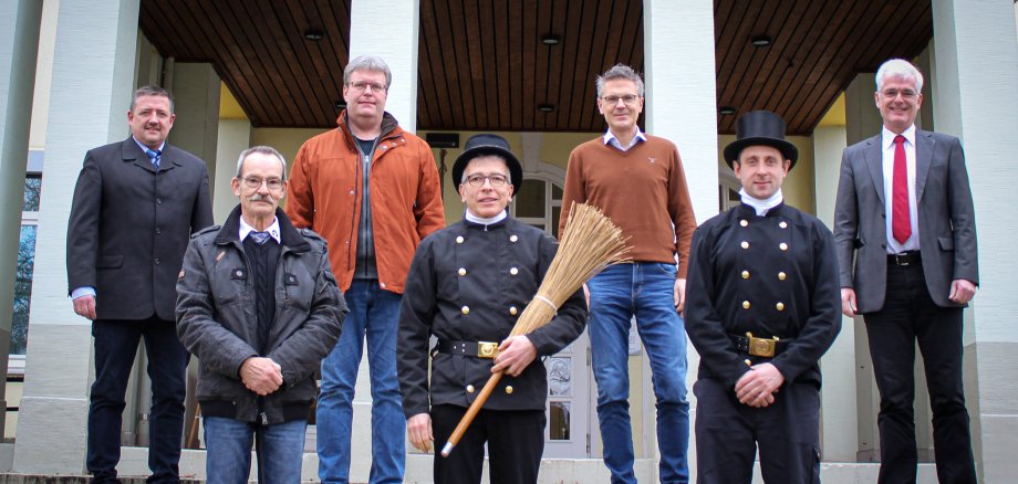 Sieben Männer, davon zwei Schornsteinfeger mit einem Besen stehen für ein Gruppenbild auf einer Treppe.
