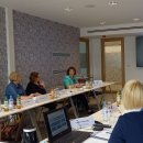 Mehrere Frauen sitzen gemeinsam in einem Besprechungsraum und unterhalten sich. An einem Fernseher an der Wand läuft eine Präsentation.