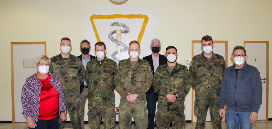 Gruppenbild mit Mundschutzmasken: Fünf Bundeswehrsoldaten stehen mit der Gesundheitsamtsleiterin und ihrem Stellvertreter sowie mit dem Landrat und seinem Stellvertreter vor einer pastellgelben Wand mit dem Gesundheitssymbol.