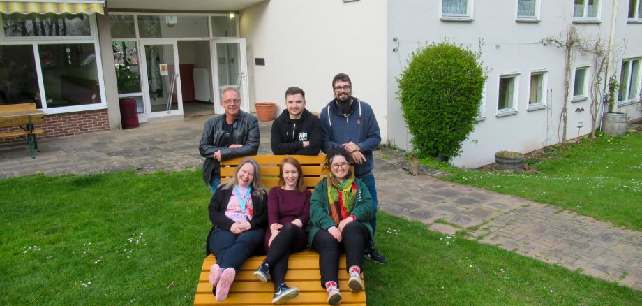 Drei Frauen liegen auf einer Holzliege, drei Männer stehen dahinter, an die Lehne gelehnt. Die Holzliege steht auf Gras, im Hintergrund ein Gebäude.