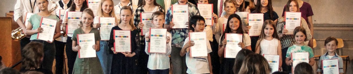 Schülerinnen und Schüler der Musikschule Hersfeld-Rotenburg stehen auf einer Bühne. Sie halten eine Urkunde. Einige haben Instrumente. Im Hintergrund ist eine Orgel. Im Vordergrund sieht man einige Gäste sitzen. Im Hintergrund hängt ein Banner der Musikschule.