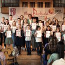 Schülerinnen und Schüler der Musikschule Hersfeld-Rotenburg stehen auf einer Bühne. Sie halten eine Urkunde. Einige haben Instrumente. Im Hintergrund ist eine Orgel. Im Vordergrund sieht man einige Gäste sitzen. Im Hintergrund hängt ein Banner der Musikschule.
