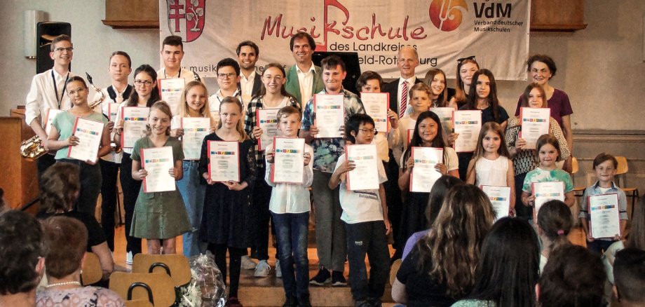 Schülerinnen und Schüler der Musikschule Hersfeld-Rotenburg stehen auf einer Bühne. Sie halten eine Urkunde. Einige haben Instrumente. Im Hintergrund ist eine Orgel. Im Vordergrund sieht man einige Gäste sitzen. Im Hintergrund hängt ein Banner der Musikschule.