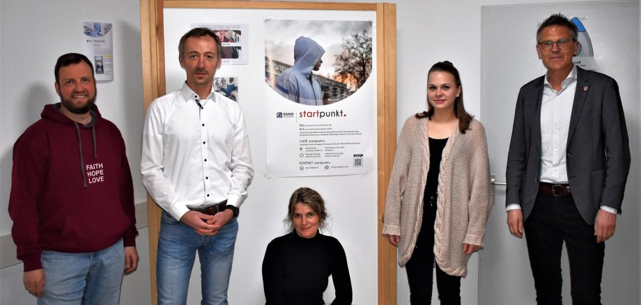 Vier Personen stehen und eine Frau hockt vor einem Holzaufsteller, an dem ein Plakat hängt mit der Überschrift "startpunkt".