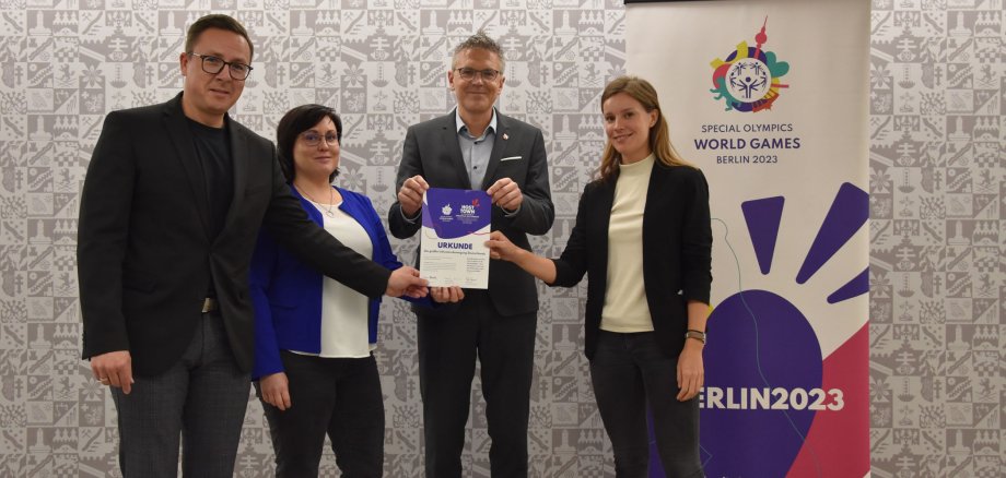 Zwei Frauen und zwei Männer zeigen eine Urkunde in die Kamera. Sie stehen neben einem Banner für die Special Olympics und vor einer Wappentapete im Landratsamt.