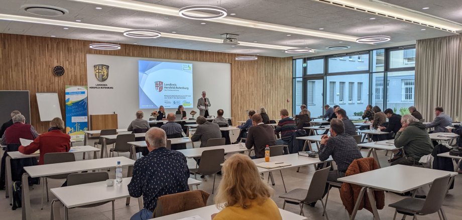 Mehrere Personen sitzen an Tischen im Kreistagssitzungssaal der Kreisverwaltung. An der Wand wird eine Präsentation gezeigt. Landrat Warnecke hält ein Mikrofon und erzählt etwas.
