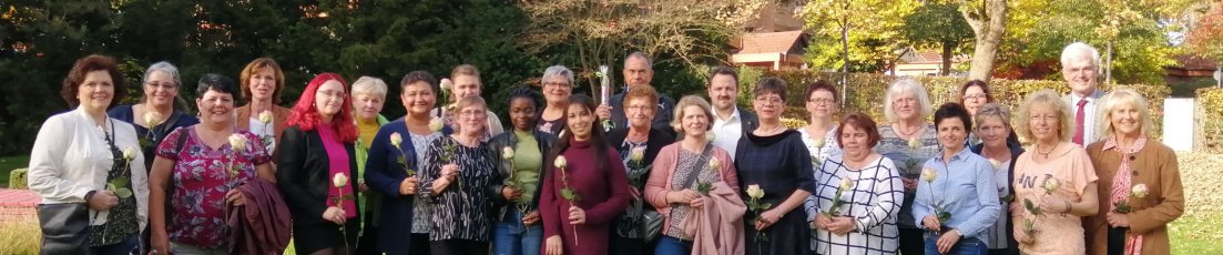 Einige Frauen und wenige Männer stehen in einer Gruppe. Die Frauen haben Rosen in der Hand, die Männer eine Stracke.