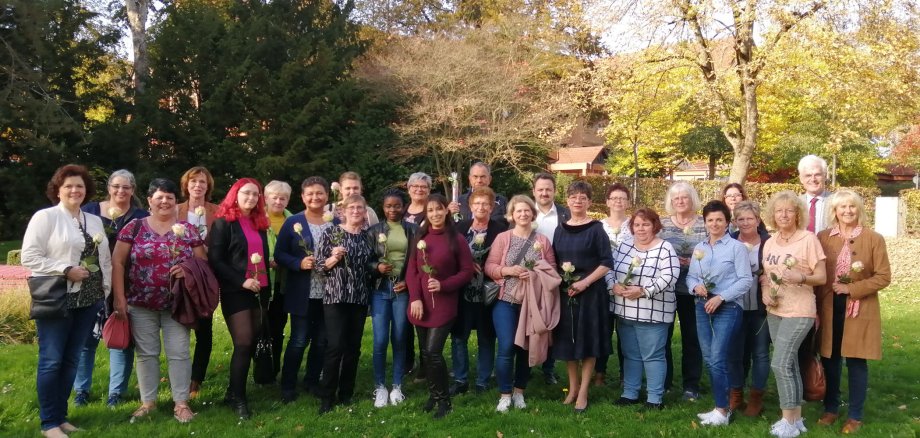 Einige Frauen und wenige Männer stehen in einer Gruppe. Die Frauen haben Rosen in der Hand, die Männer eine Stracke.