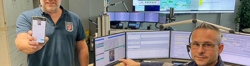 Der Leitstellenchef steht und einer seiner Mitarbeiter sitzt an seinem Arbeitsplatz in der Zentralen Leitstelle der Kreisverwaltung. Die beiden präsentieren ein Handy. Im Hintergrund sieht man den Arbeitsplatz des Leitstellenmitarbeiters.