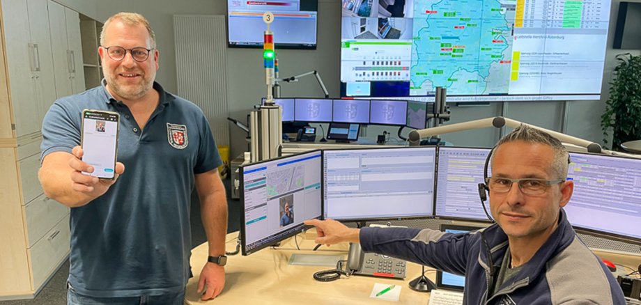 Der Leitstellenchef steht und einer seiner Mitarbeiter sitzt an seinem Arbeitsplatz in der Zentralen Leitstelle der Kreisverwaltung. Die beiden präsentieren ein Handy. Im Hintergrund sieht man den Arbeitsplatz des Leitstellenmitarbeiters.