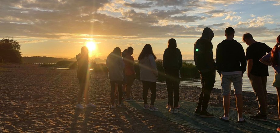 Eine Gruppe Jugendlicher steht an einem Sandstrand. Die Sonne geht gerade unter, der Himmel ist orange-blau. Es sind ein paar Wolken.