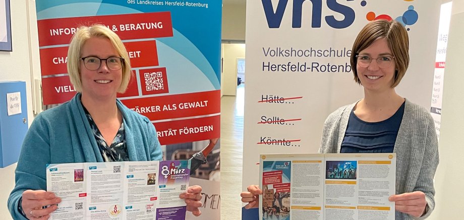 Die Frauenbeauftragte und eine VHS-Mitarbeiterin halten ein Schild mit Werbung zur Kooperation der beiden Organisationen in der Hand.