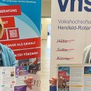 Die Frauenbeauftragte und eine VHS-Mitarbeiterin halten ein Schild mit Werbung zur Kooperation der beiden Organisationen in der Hand.