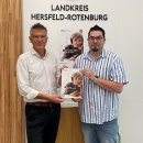 Zwei männliche Personen stehen in einem Sitzungsraum vor einer Wand mit dem Schriftzug "Landkreis Hersfeld-Rotenburg". Sie halten ein Programmheft der Volkshochschule Hersfeld-Rotenburg in der Hand.