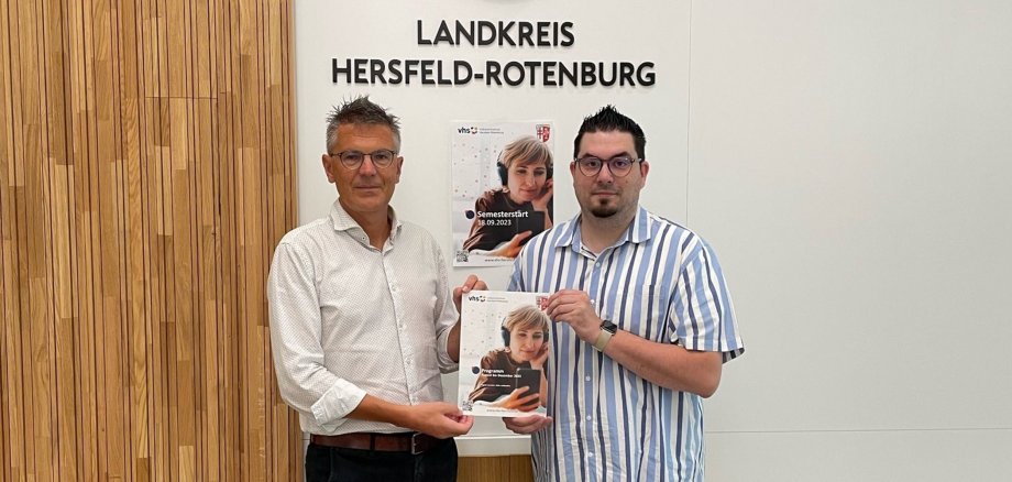 Zwei männliche Personen stehen in einem Sitzungsraum vor einer Wand mit dem Schriftzug "Landkreis Hersfeld-Rotenburg". Sie halten ein Programmheft der Volkshochschule Hersfeld-Rotenburg in der Hand.