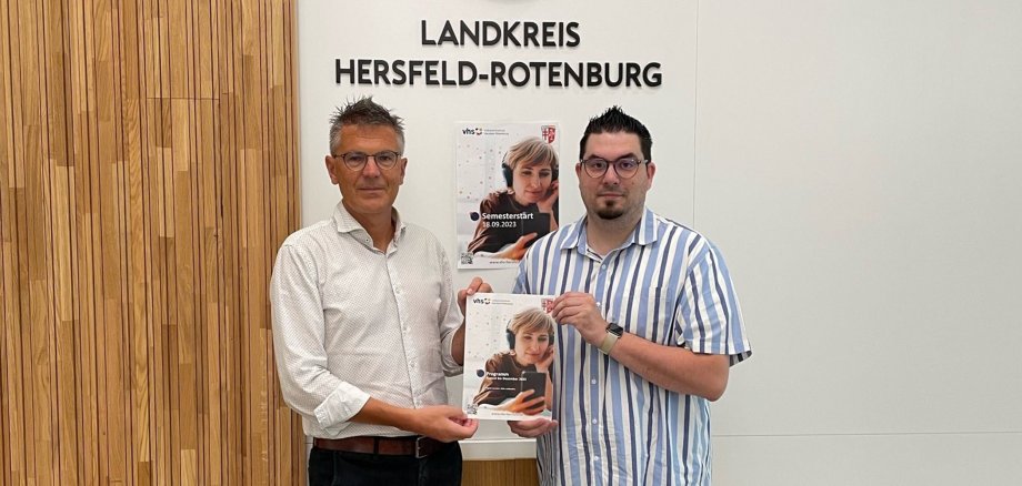 Zwei männliche Personen stehen in einem Sitzungsraum vor einer Wand mit dem Schriftzug "Landkreis Hersfeld-Rotenburg". Sie halten ein Programmheft der Volkshochschule Hersfeld-Rotenburg in der Hand.