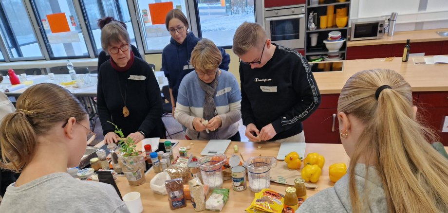 Zwei ältere Personen und mehrere Jugendliche stehen in einer Küche in einer Schule. Sie haben viele Lebensmittel in der Küche und kochen gemeinsam.