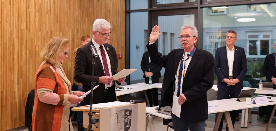 Drei Personen stehen im Vordergrund. Die beiden linken Personen lesen etwas auf einem Blatt. Die Person, die ganz rechts steht hat die Hand gehoben und legt einen Eid ab. Im Hintergrund beobachten weitere Männer die Szene.