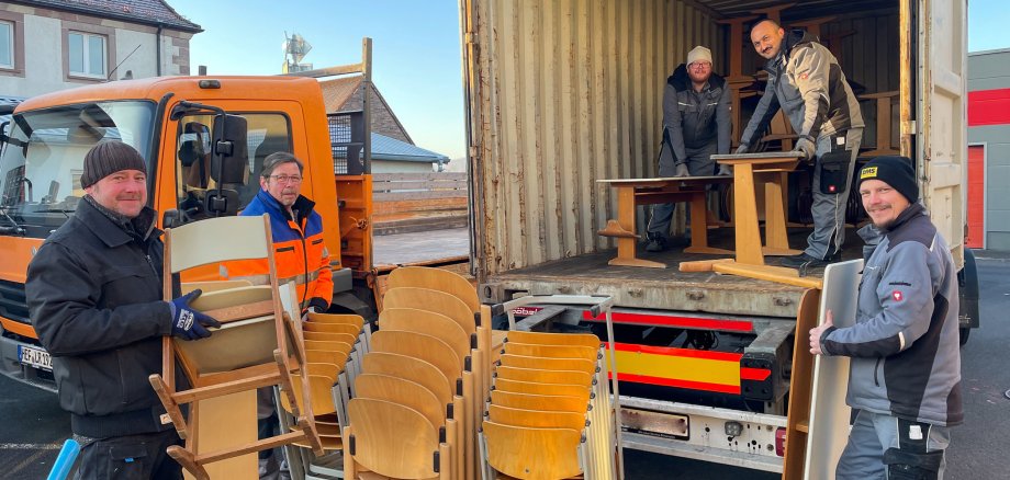 Drei Männer stehen mit alten Schulstühlen und Schultischen in der Hand vor einem LKW-Container, in dem zwei weitere Männer stehen, die gerade Schultische im Container stapeln. Im Hintergrund ist ein weiteres Auto mit Ladefläche zu sehen.