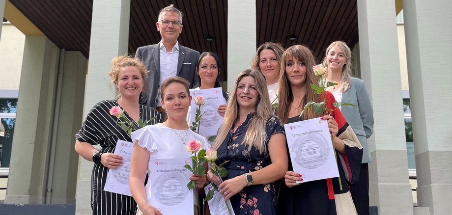 Gruppenbild auf der Treppe vor dem Hersfelder Landratsamt: Der Erste Kreisbeigeordnete Dirk Noll und Isabell Schmidt von der Fachberatung Kindertagespflege beglückwünschen (hinten rechts) sechs neue Tagesmütter im Landkreis Hersfeld-Rotenburg.  Die Tagesmütter zeigen ihre Teilnahmebescheinigung und geschenkte rosa Rose.