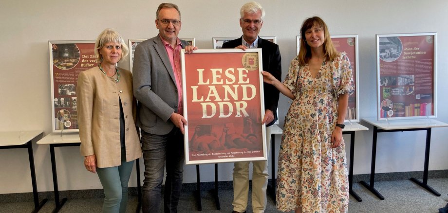 Vier Personen darunter Landrat Torsten Warnecke stehen in einer Reihe in der Modellschule Obersberg. Gemeinsam halten sie ein Poster, das Teil der Ausstellung "Leseland DDR" ist. Im Hintergrund sieht man weitere Teile der Ausstellung, die in der Modellschule Obersberg eröffnet wurde.