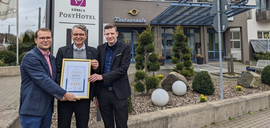 Drei Personen halten ein gerahmtes Zertifikat in den Händen. Sie stehen vor dem Rotenburger Posthotel.