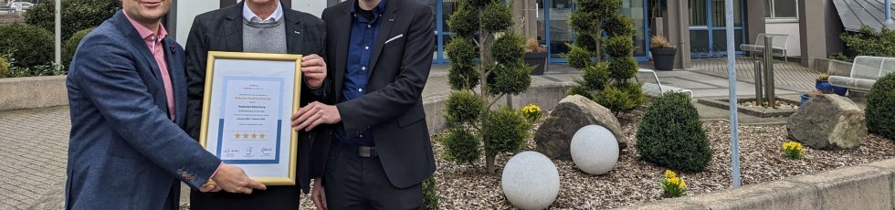 Drei Personen halten ein gerahmtes Zertifikat in den Händen. Sie stehen vor dem Rotenburger Posthotel.