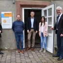 Vier Männer und eine Frau stehen vor einem Gebäude mit offener Tür. Am Gebäude hängt ein Schild, auf dem das Projekt A³ abgebildet ist.